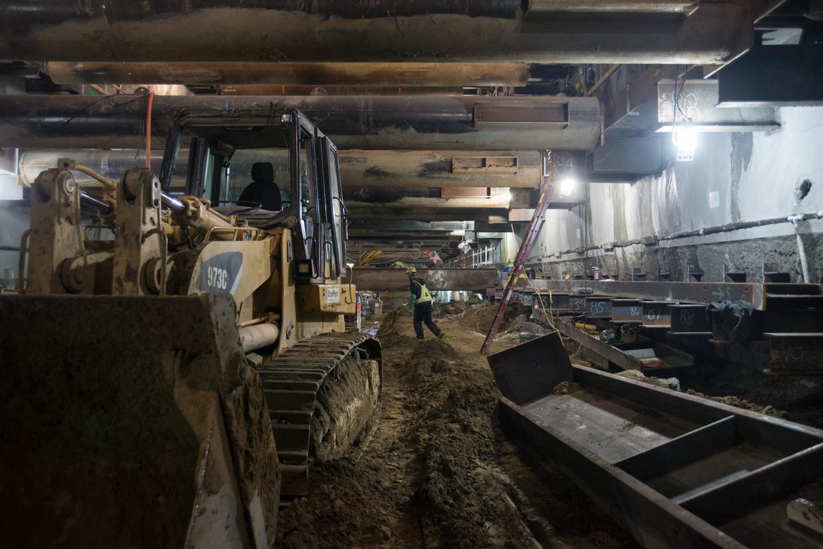 Skid steer loader operating in underground station excavation with workers and temporary bracing visible.