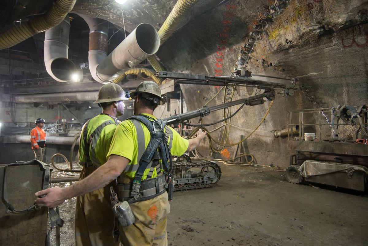 Construction workers in safety gear operating drilling equipment in underground tunnel during transit station excavation work.