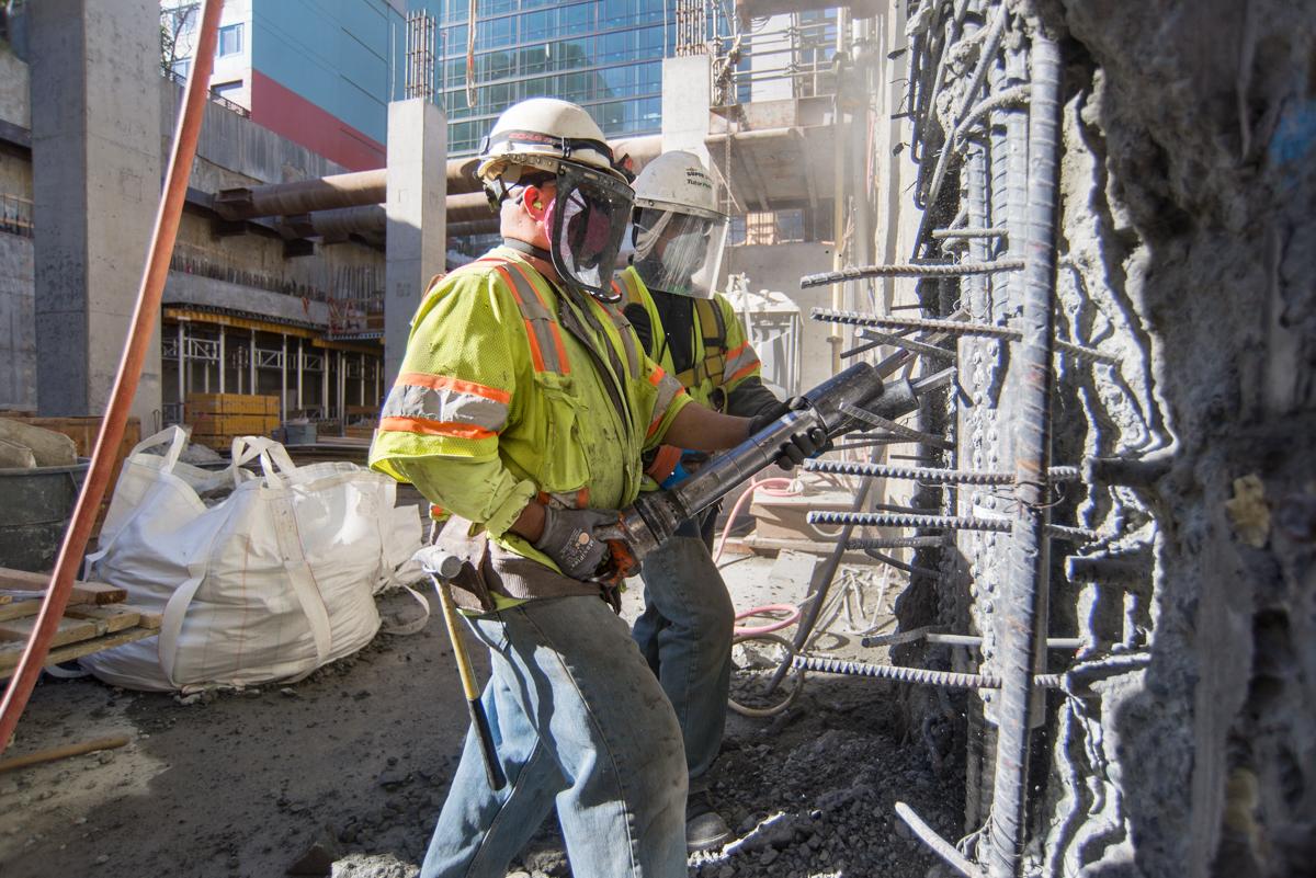 Construction worker in safety gear operating pneumatic drill on concrete wall at urban construction site.