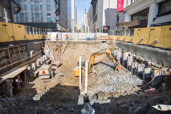 Construction excavation site on urban street with yellow barriers, heavy equipment, and surrounding buildings.