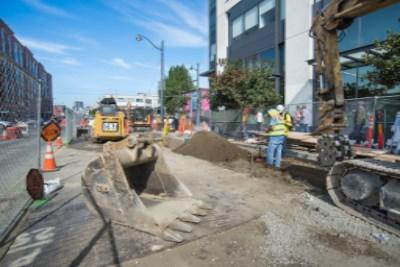Construction crew installing new sewer line in urban trench on 4th Street with equipment and safety barriers.