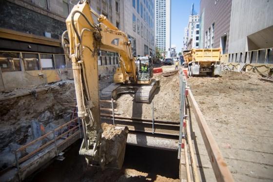 Yellow excavator on Stockton Street construction site with trucks and urban buildings in background.