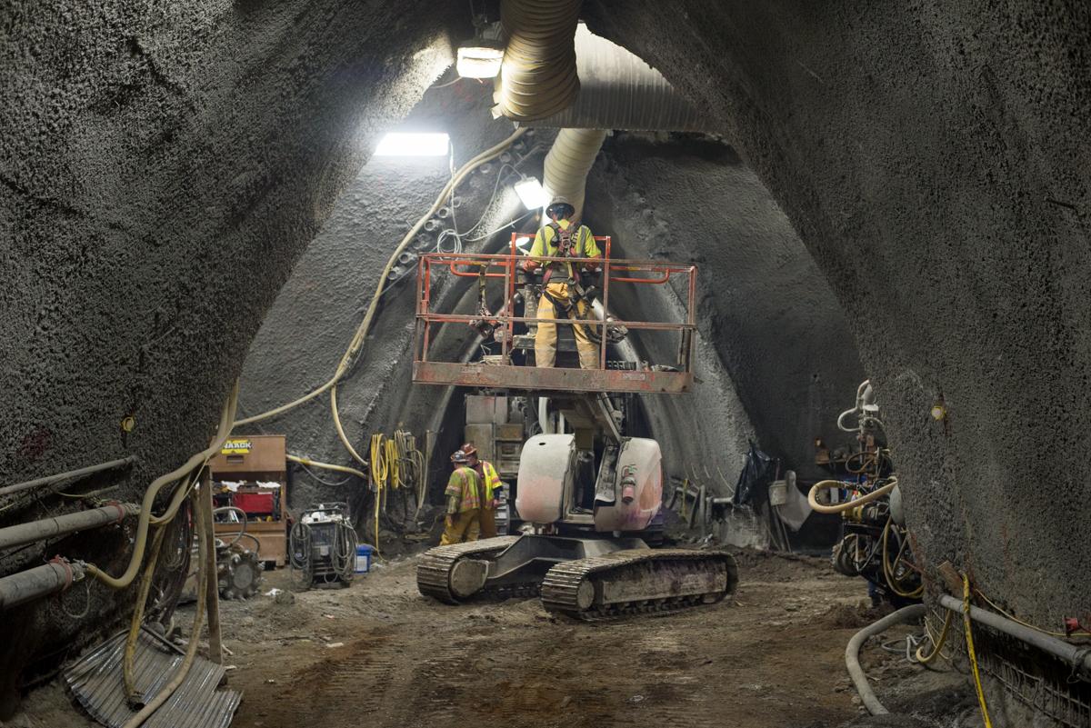 Workers on elevated platform operate equipment inside underground transit tunnel cavern during construction.