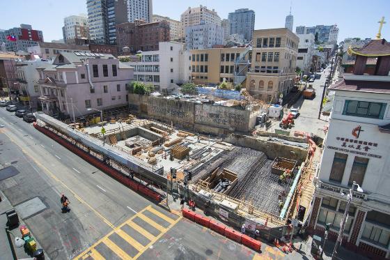 Aerial view of Chinatown Station construction site with red barriers, exposed foundation, and surrounding buildings.