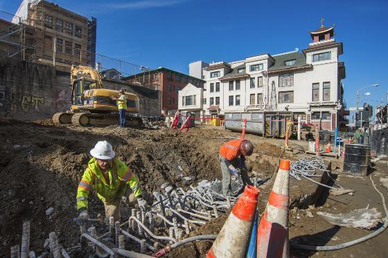 Workers in safety gear remove rebar from an urban construction site surrounded by buildings.