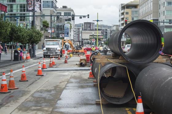 Large black sewer pipes staged on 4th Street with construction equipment and traffic cones during Central Subway project work.