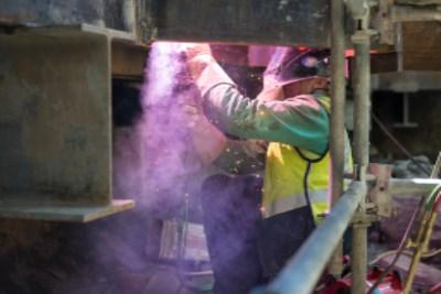 Worker welding steel beam inside transit headhouse during construction, purple welding light visible.