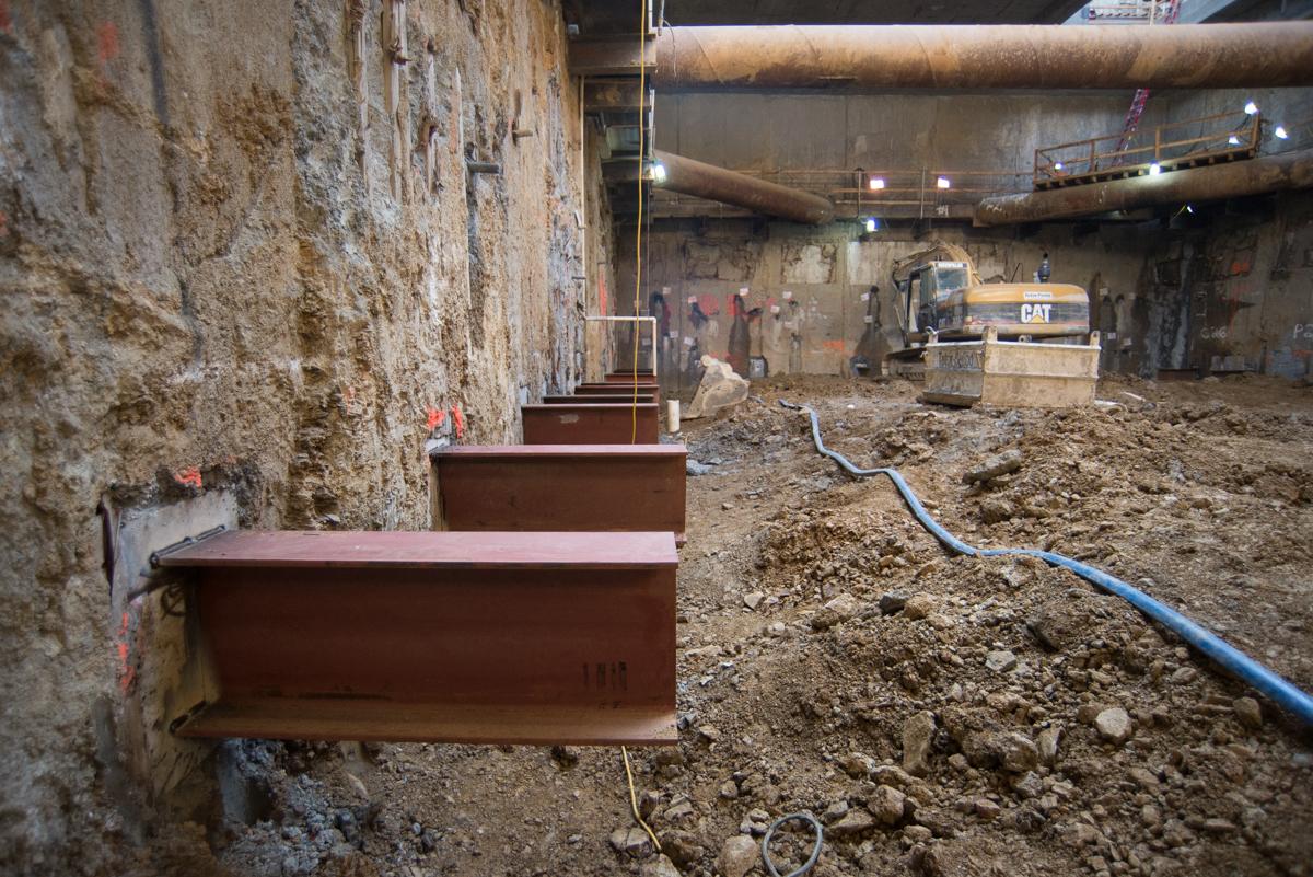Steel I-beams installed along slurry wall at Chinatown Station construction site with excavator and workers visible.