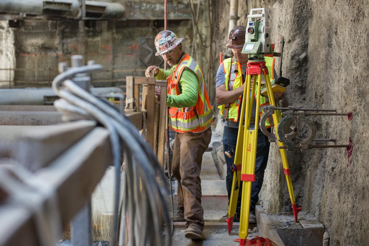 Construction workers use surveying equipment on a narrow catwalk against a concrete wall at a transit station site.