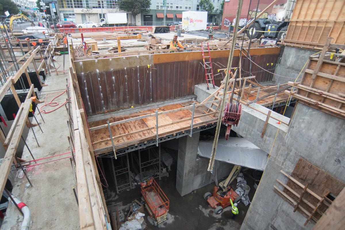 Construction site with excavation pit, concrete forms, steel shoring, and workers preparing foundation work for future station.