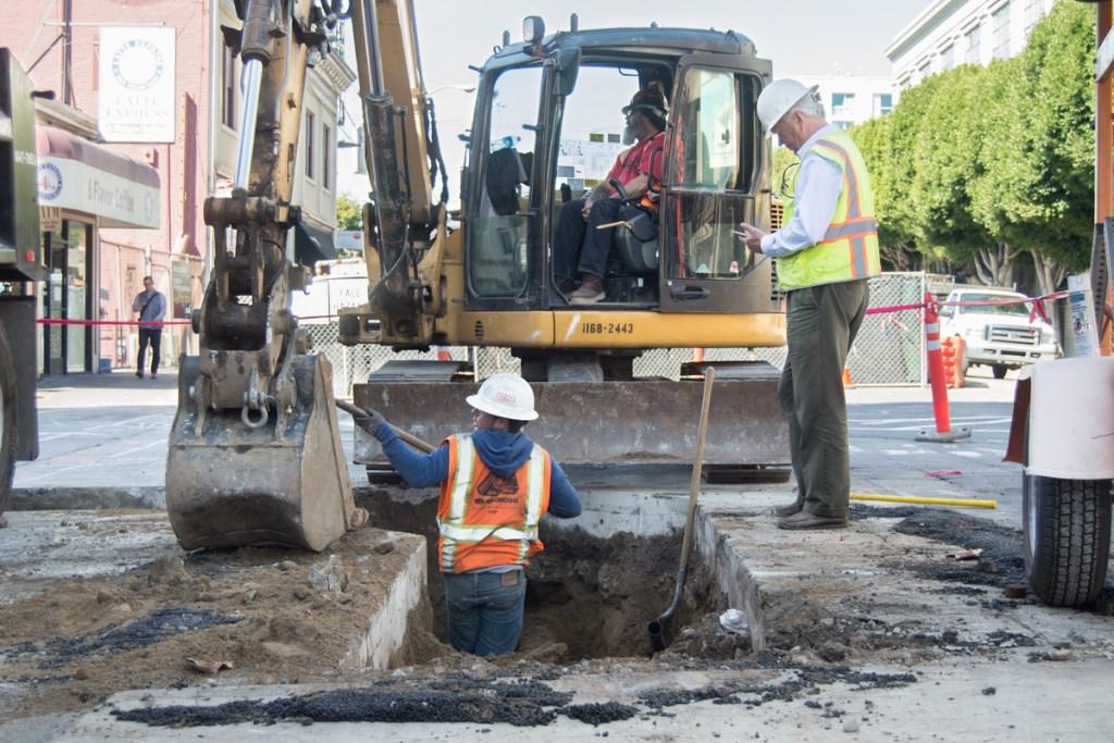 Construction crew excavates underground utilities on city street with backhoe and workers in safety gear.