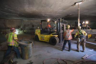 Workers in safety vests operate a forklift in an underground transit station under construction with temporary support structures.