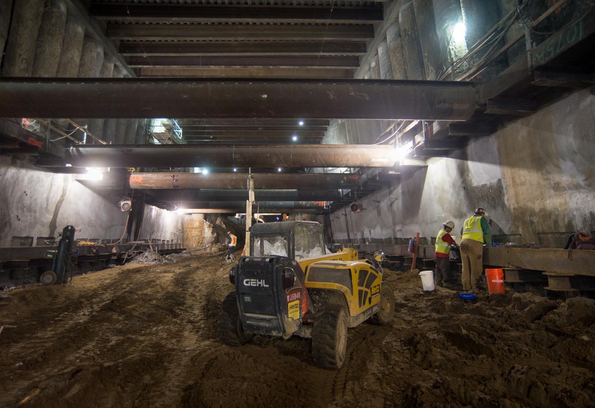 Underground station excavation with heavy equipment and workers in temporary bracing area beneath city streets.