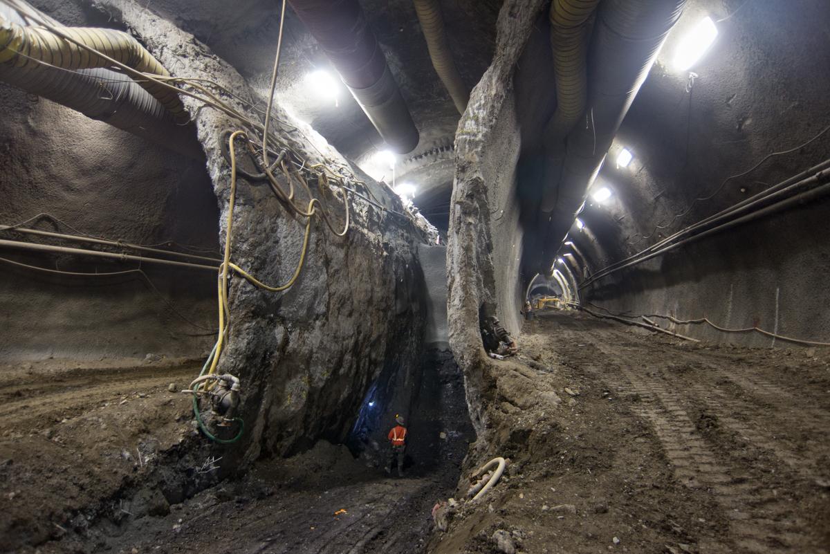 Worker in safety vest stands beneath large concrete support pillar in underground transit tunnel with LED lighting.