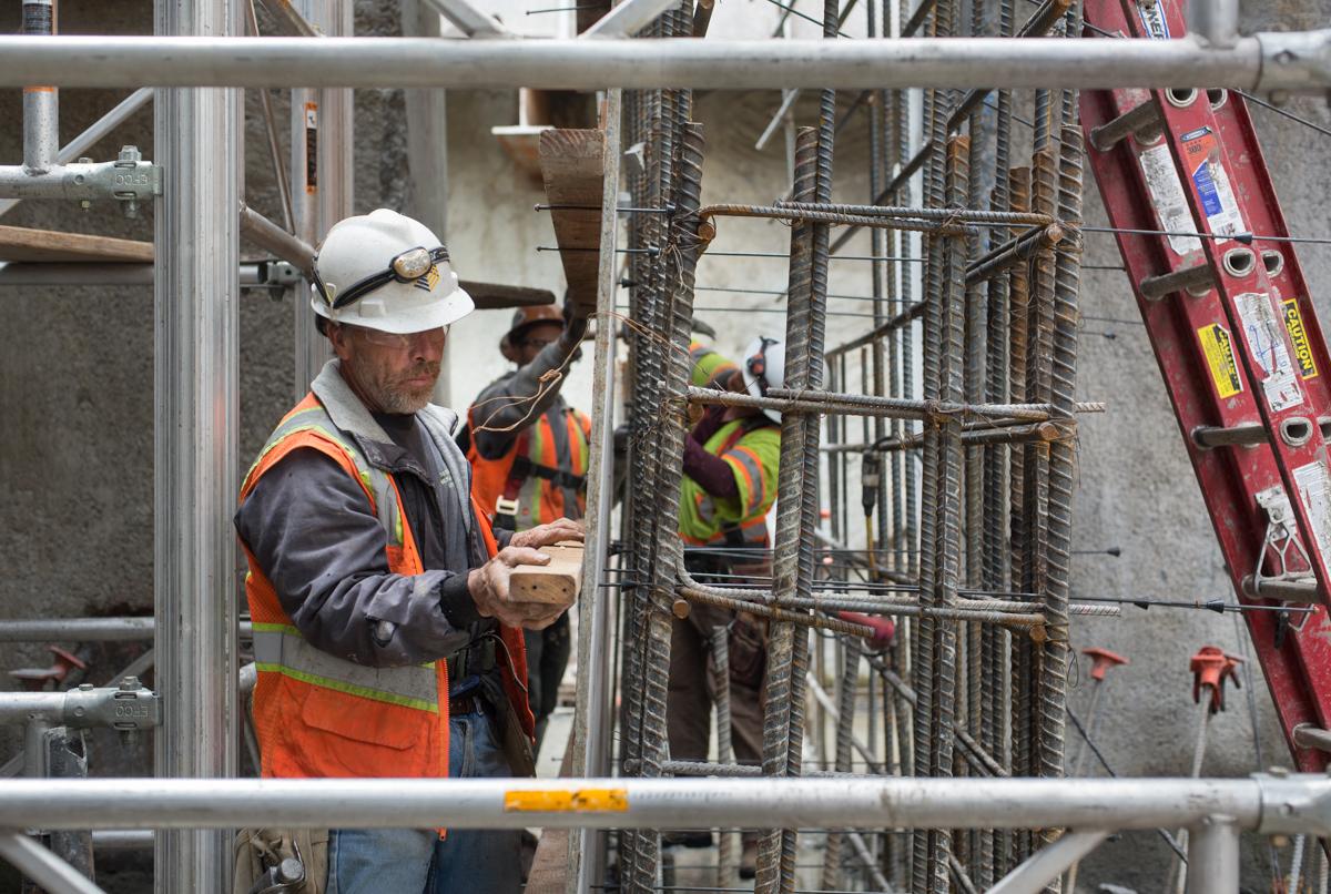 Construction worker in hard hat and safety vest assembles concrete forms on mezzanine level near rebar cage.
