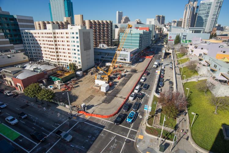 Construction site on 4th Street with cranes, equipment, and orange barriers; city skyline in background.