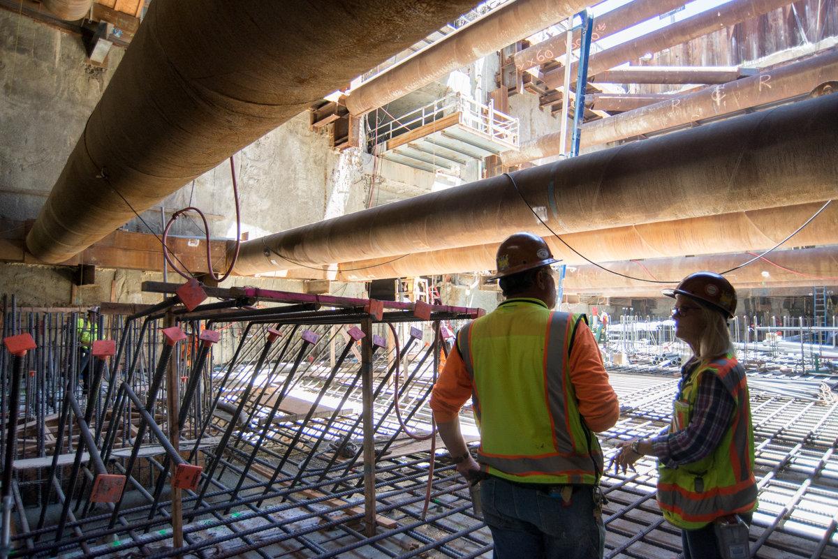 Construction workers in safety vests inspect rebar cage assembly under concrete bridge structure at transit construction site.