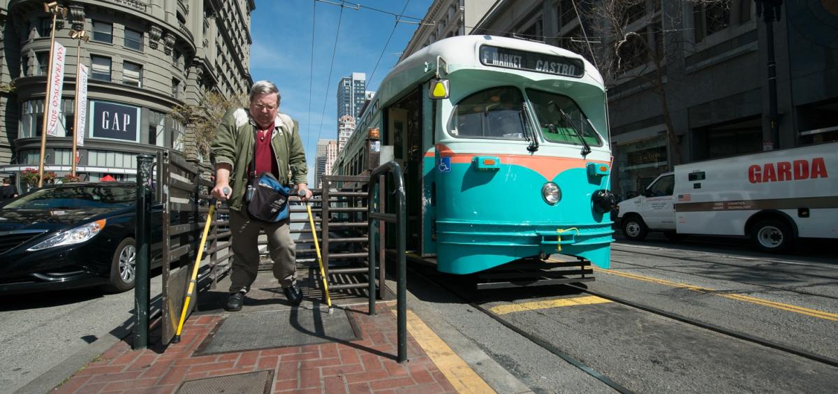 Man using wheelchair lift to board turquoise and white streetcar on Market Street in San Francisco.