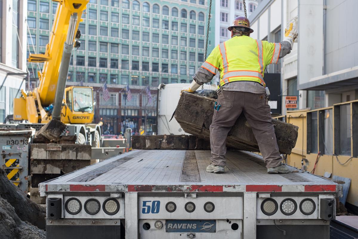 Construction worker receives wooden cribbing from crane operator on flatbed truck at downtown transit worksite.
