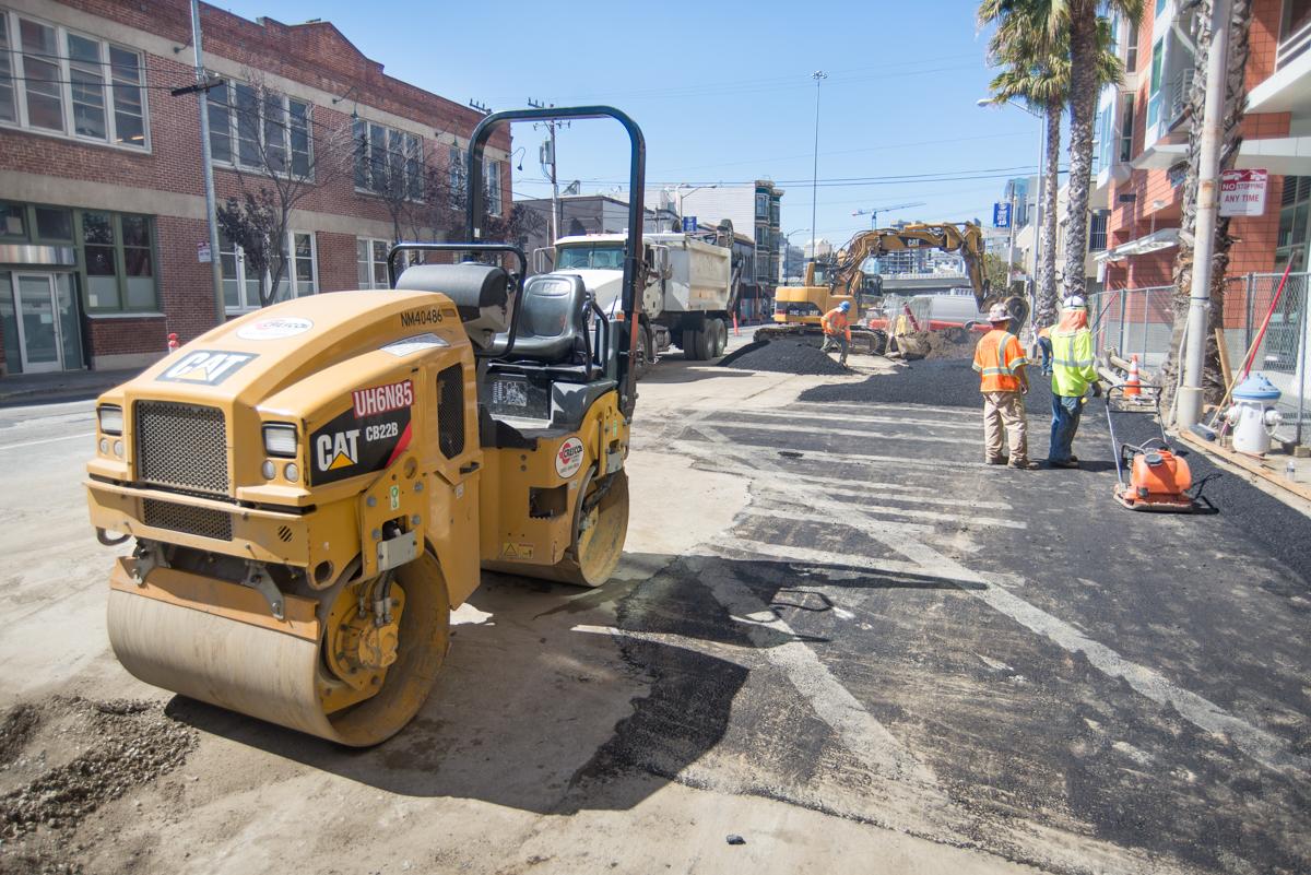 Road construction crew operating a yellow CAT roller compactor on a city street during pavement work.