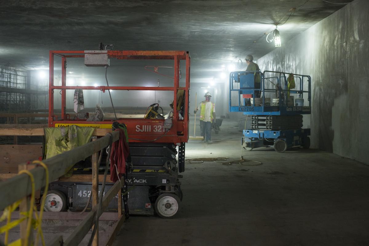 Workers operate scissor lifts and equipment inside underground station construction area with concrete walls and overhead lighting.
