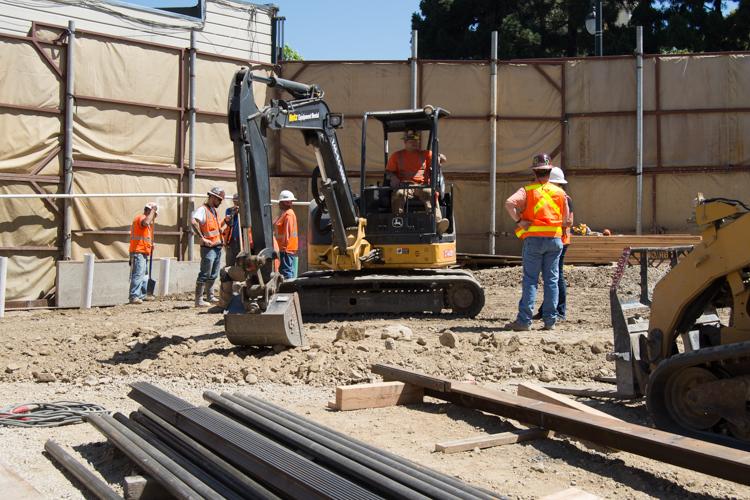 Construction workers in safety vests operate excavation equipment at a job site surrounded by protective barriers.