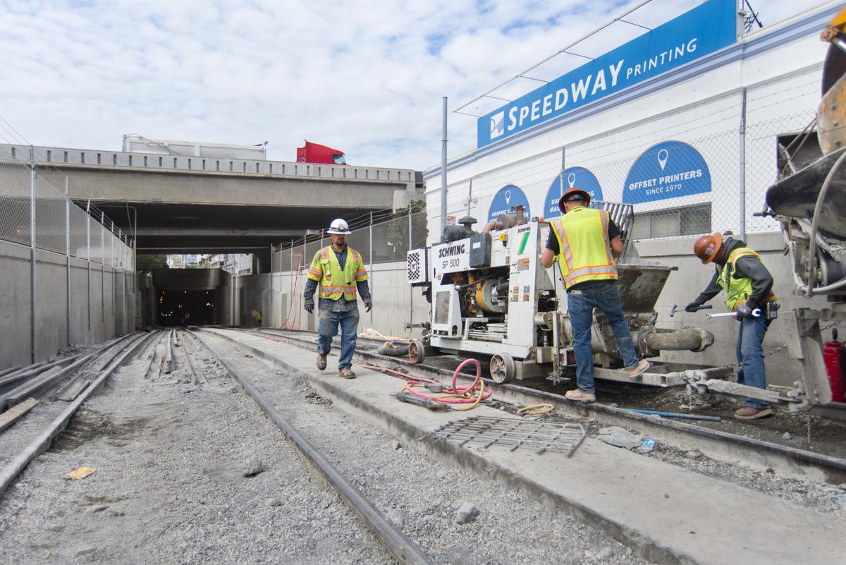 Workers in safety vests operate concrete pump equipment on transit tunnel construction site under highway overpass.