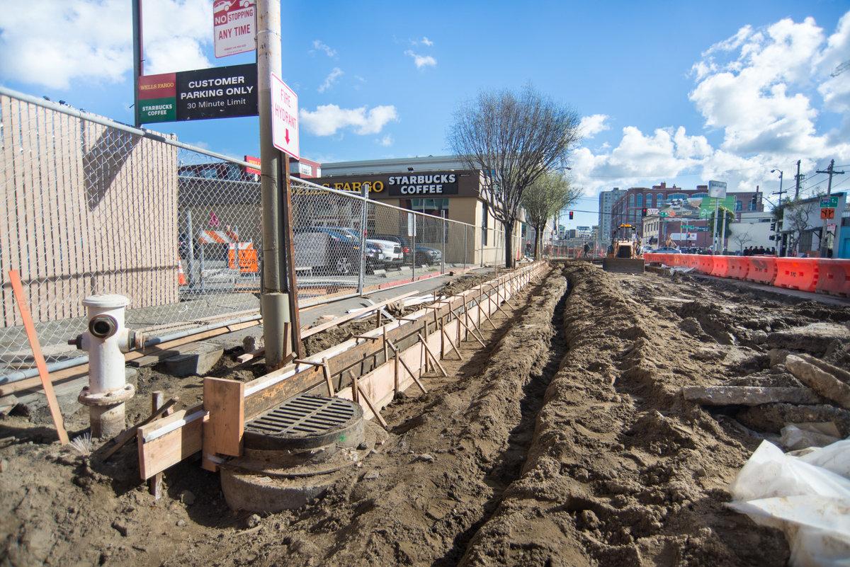 Construction site showing excavated trench, concrete forms, and Starbucks storefront along 4th Street for Central Subway project.