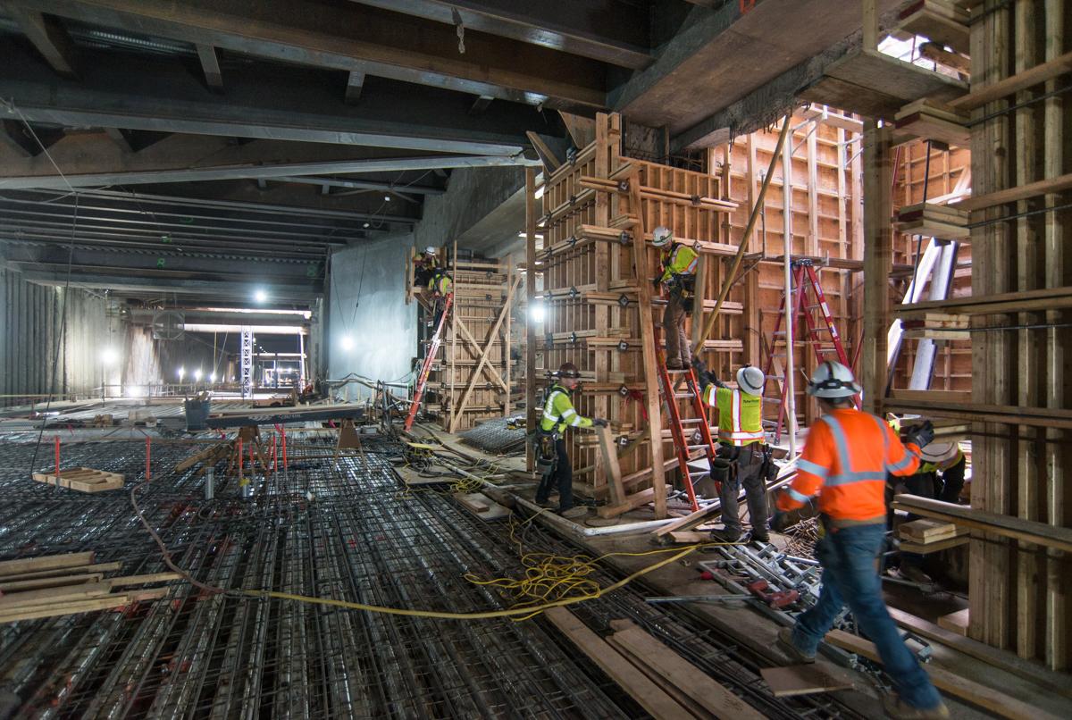 Workers in safety vests construct wooden forms and scaffolding for concrete columns inside a transit station concourse under construction.