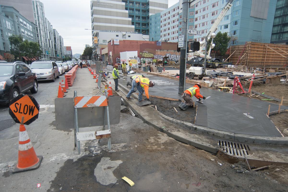 Construction work on 4th Street with orange cones, barriers, and workers in safety vests on an active job site.