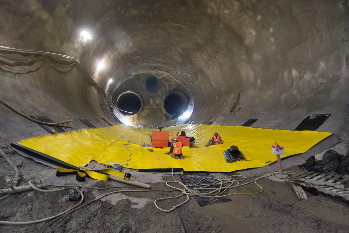 Workers in safety gear on yellow platform inside large concrete tunnel with circular openings and overhead lighting.
