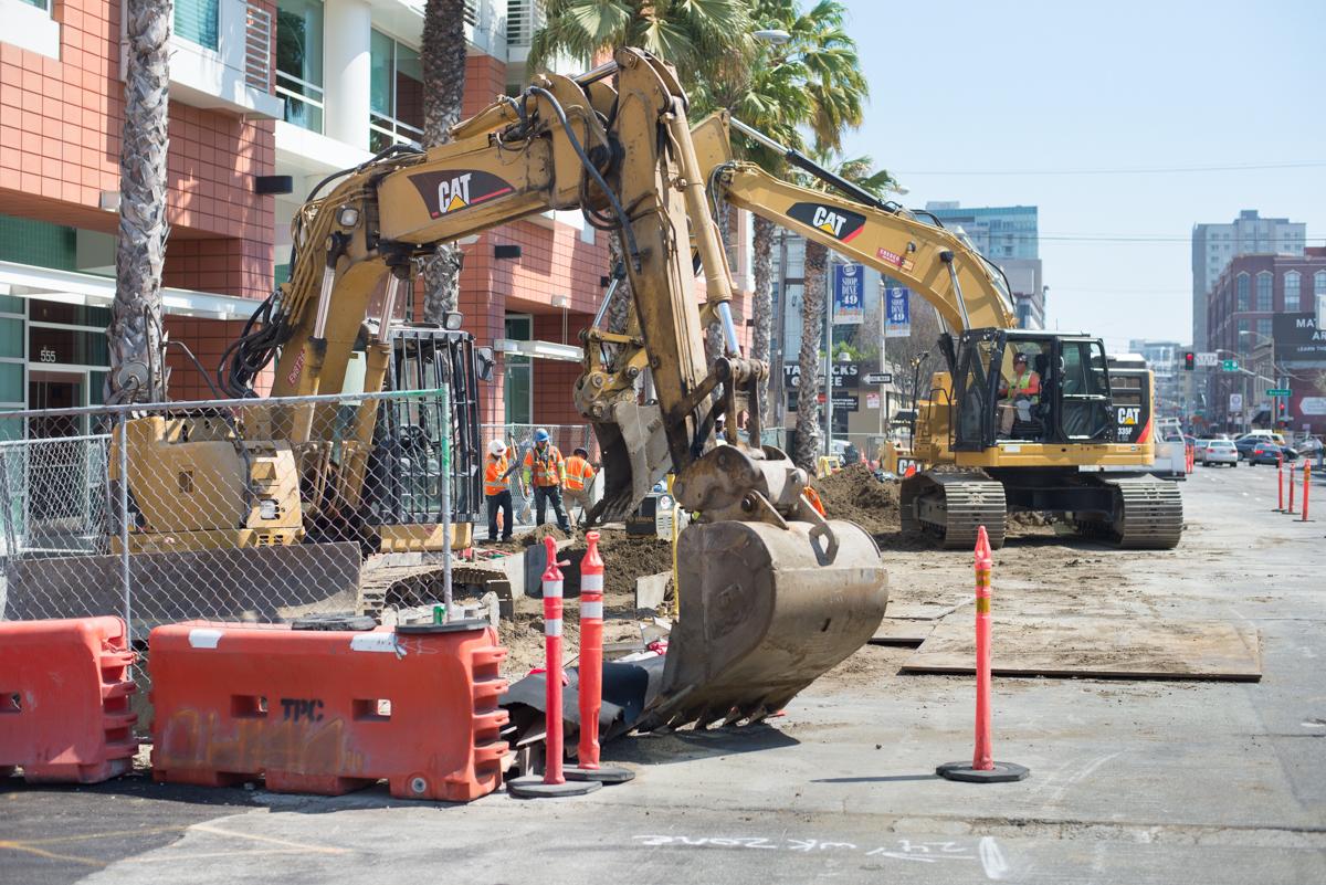 Construction excavators and workers at urban transit project site with safety barriers and traffic cones.