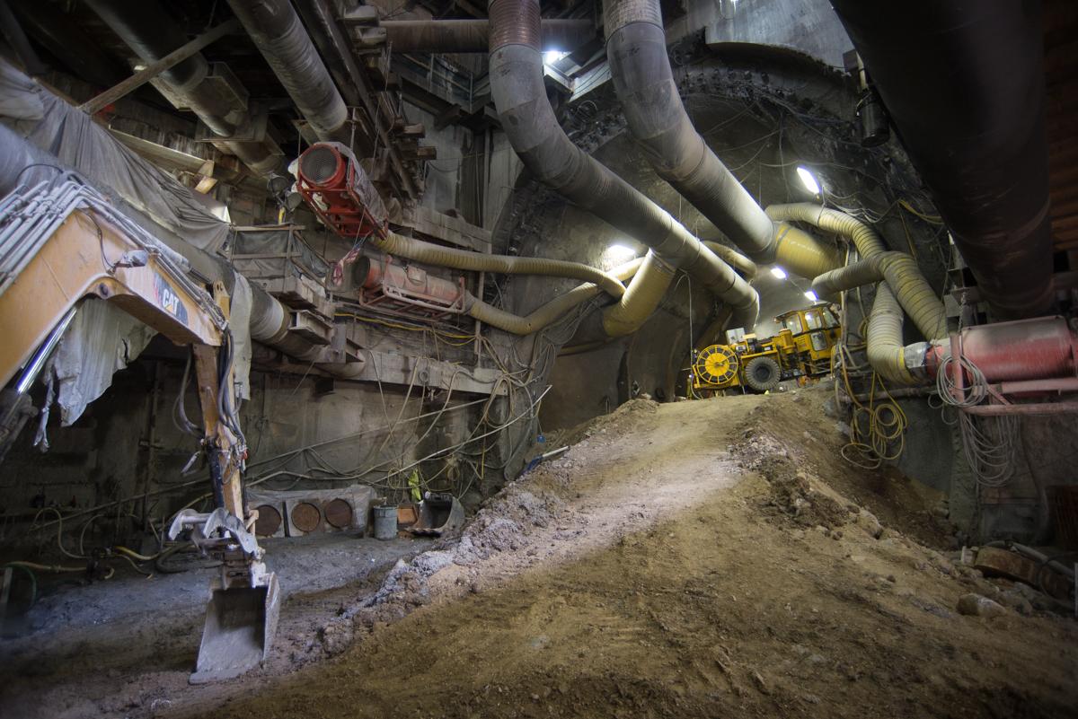 Underground tunnel construction site with large pipes, excavated earth, and yellow work lights illuminating the cavern space.