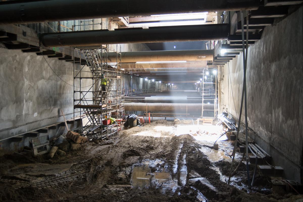 Underground tunnel construction site with steel bracing, scaffolding, and debris under city street.