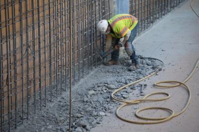 Worker in yellow safety vest using jackhammer to remove concrete at construction site with rebar fencing and hose visible.