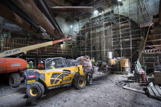 Construction site with yellow telehandler, red lift, metal scaffolding, and workers in safety gear inside industrial building.