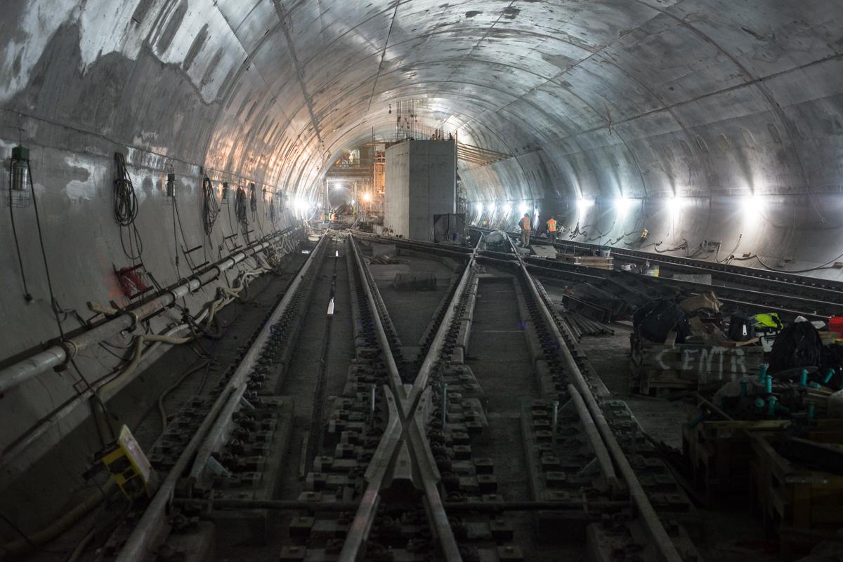 Underground tunnel with multiple railroad tracks under construction, lit by overhead lights with workers visible in the distance.