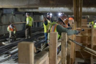 Workers in safety vests construct wooden forms inside a transit station excavation trench.