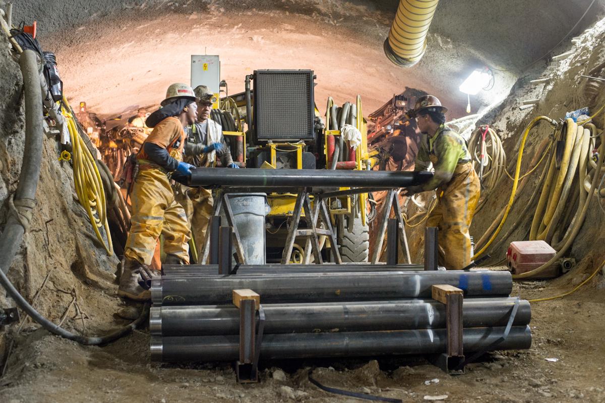Workers in hard hats and safety gear handle large black pipe sections underground during tunnel excavation and soil improvement work.