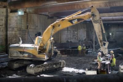 Yellow CAT excavator positioning steel I-beam during headhouse excavation work at transit station.