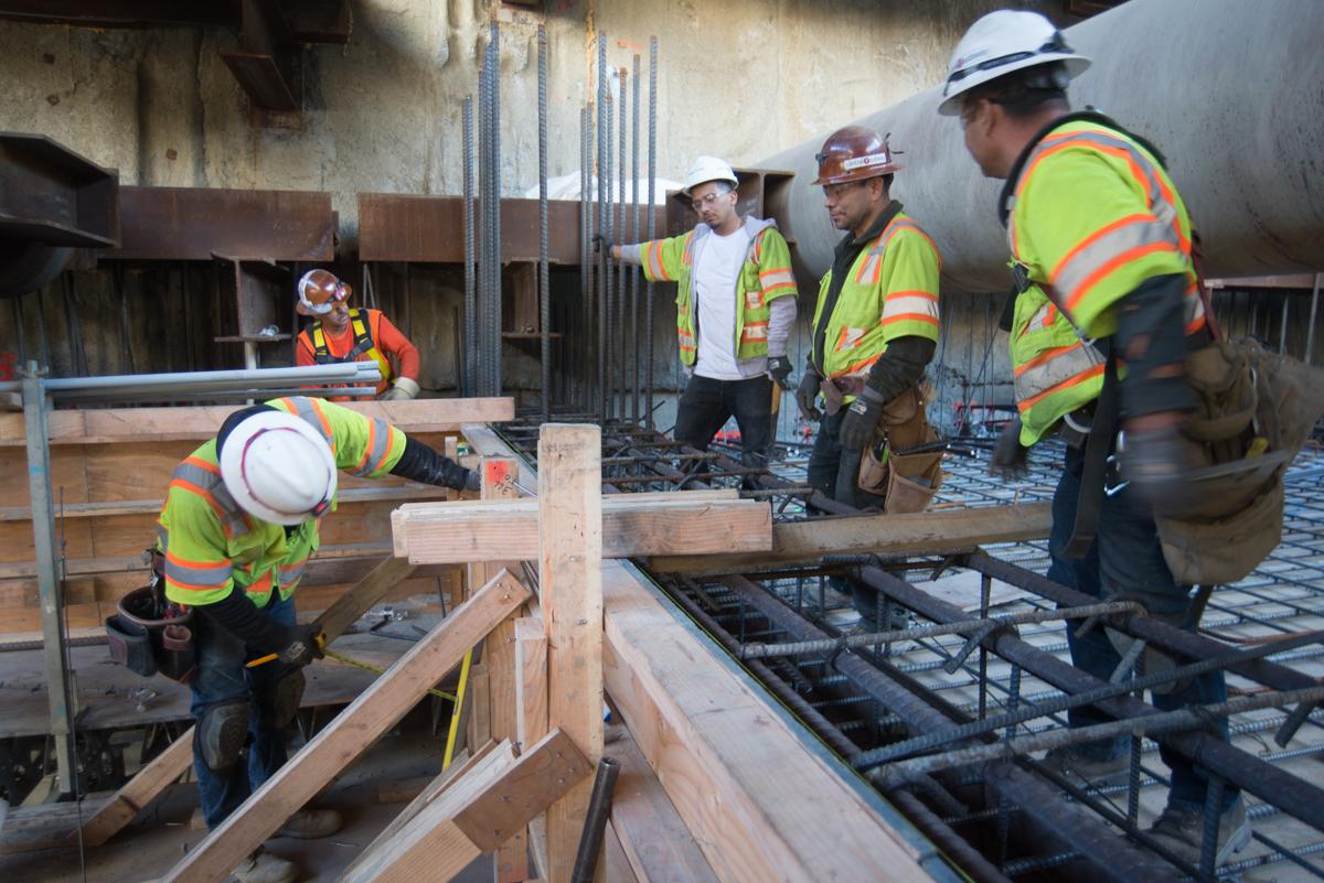 Construction workers in safety gear prepare concrete forms and rebar for mezzanine floor slab work inside a transit station.