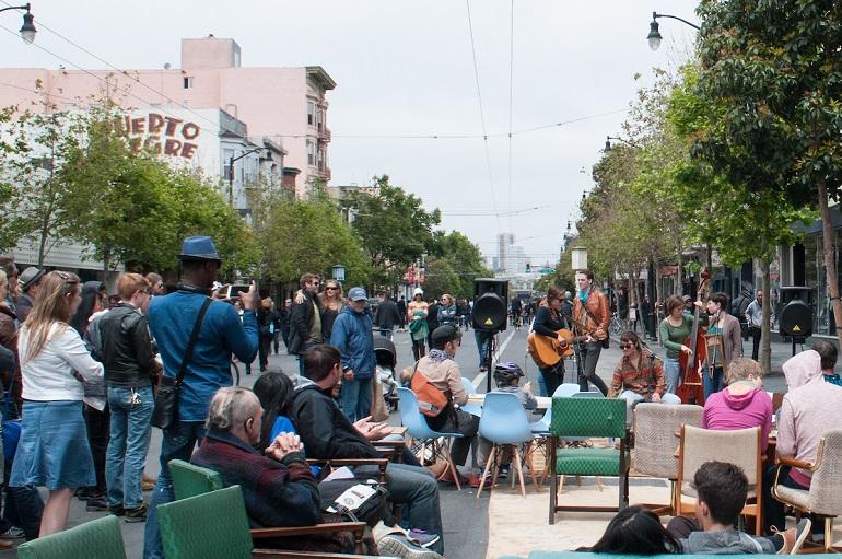 Crowded street festival with pedestrians, musicians, and seated spectators on a closed urban street.