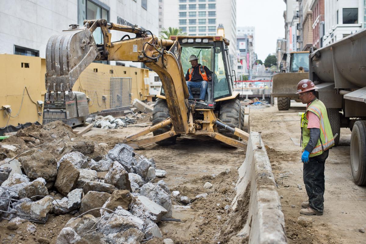 Excavator loads concrete tunnel debris into dump truck at urban construction site with worker supervising.