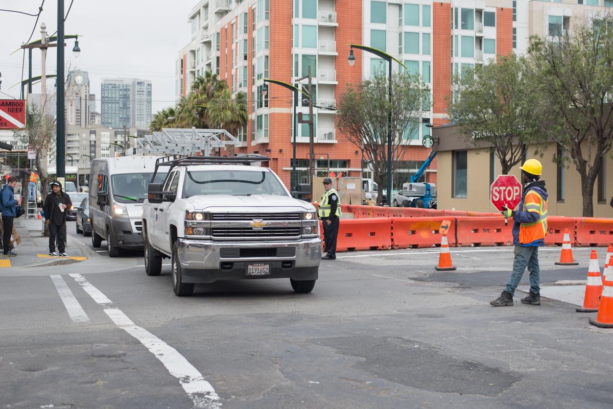 Construction crew manages traffic at street intersection with stop sign, traffic cones, and safety personnel in high-visibility gear.