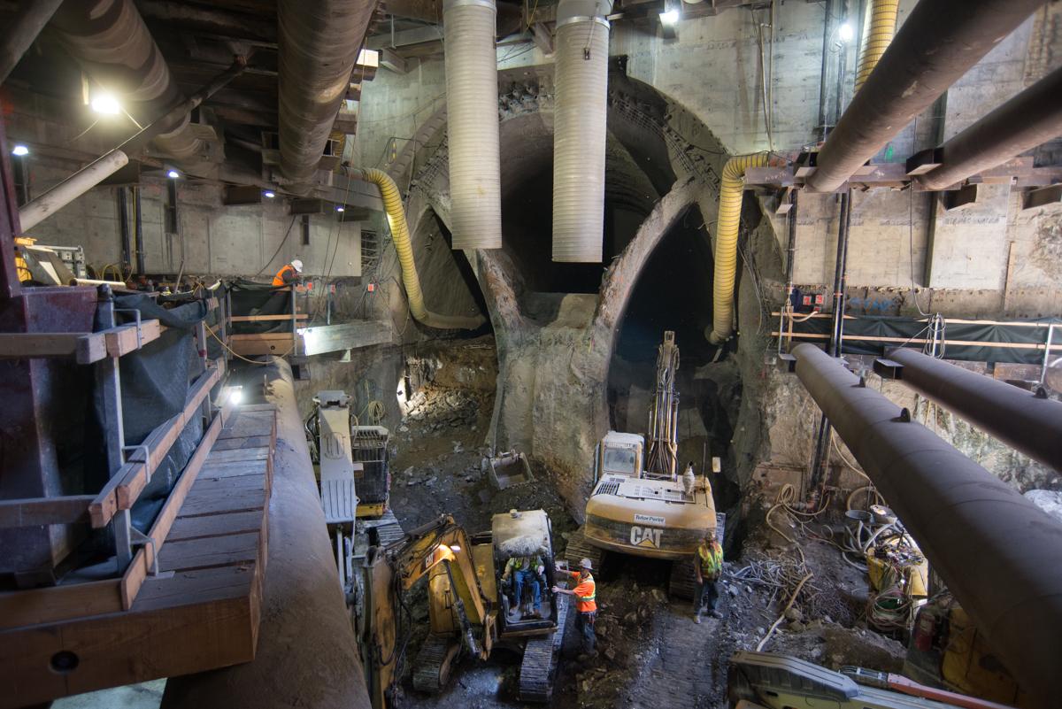 Construction equipment and workers excavate underground cavern with large pipes and support columns visible overhead.