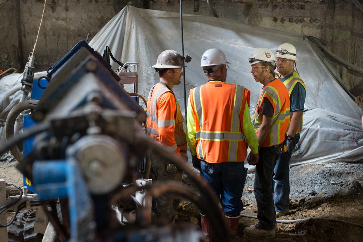 Construction workers in safety vests and hard hats confer at an underground work site with concrete walls and equipment visible.