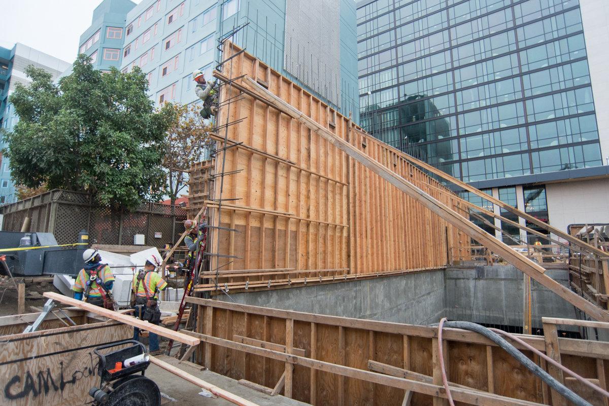 Construction workers install wooden formwork and rebar for ventilation structure along urban street.