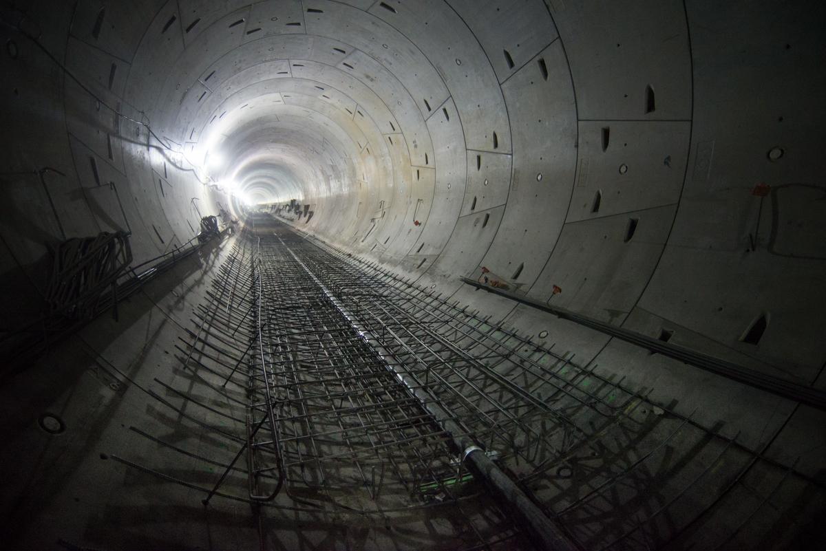 Interior view of subway tunnel under construction with concrete walls, steel reinforcement, and light at end of tunnel.