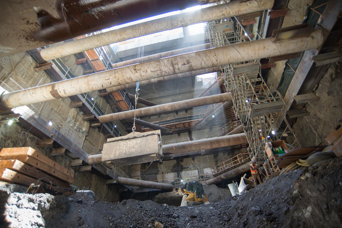 Large industrial pipes and structural beams visible in underground construction shaft with work equipment and lighting.