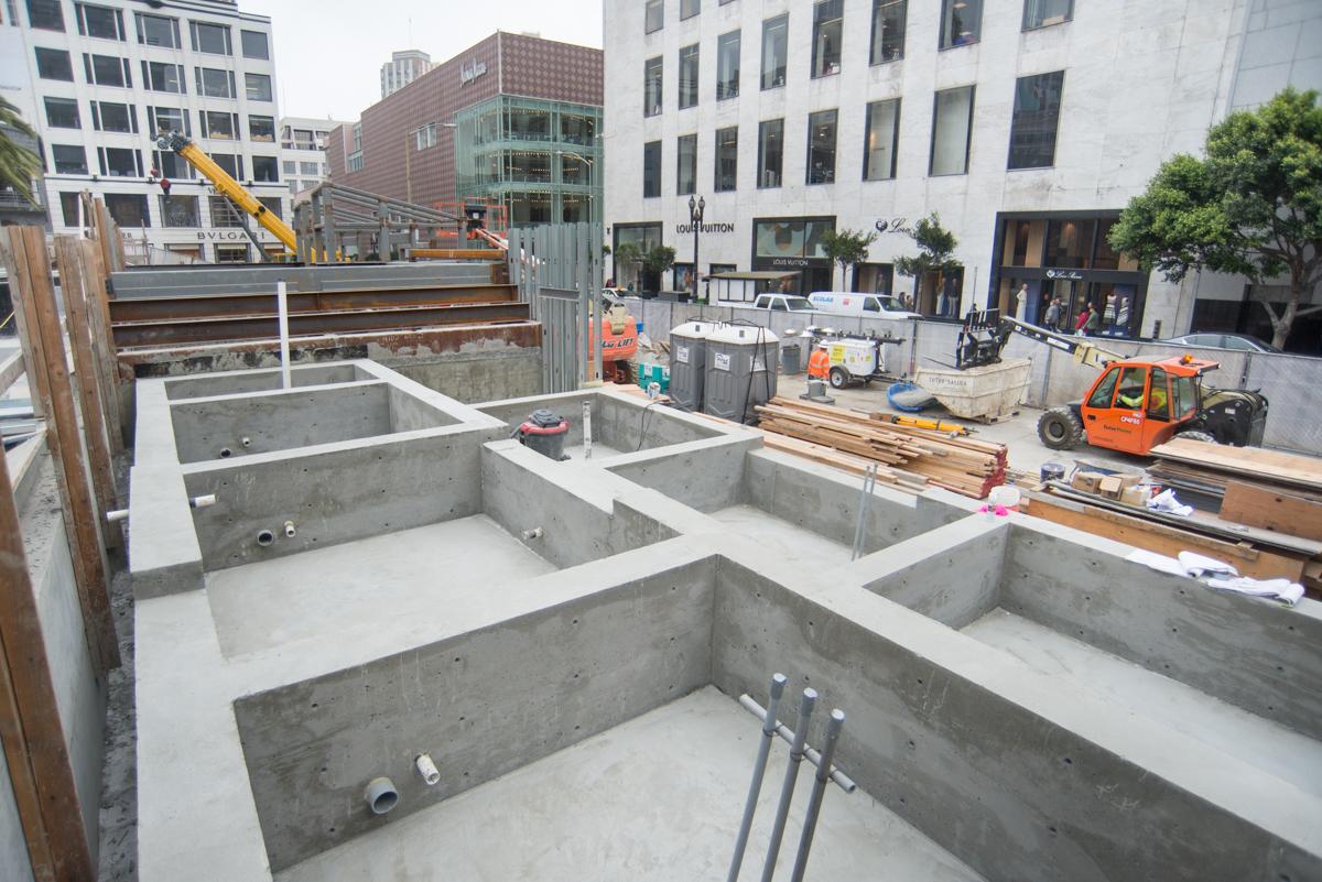 Underground construction site with concrete foundation walls and structural elements at street level in urban setting.